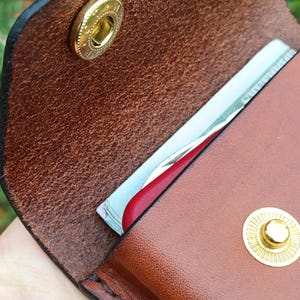 May include: A brown leather wallet featuring a textured grain, secured with two gold-toned metal snap buttons. The wallet is partially open, revealing a stack of paper currency and a red card edge tucked inside the main compartment.