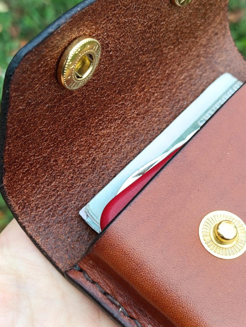 May include: A brown leather wallet featuring a textured grain, secured with two gold-toned metal snap buttons. The wallet is partially open, revealing a stack of paper currency and a red card edge tucked inside the main compartment.