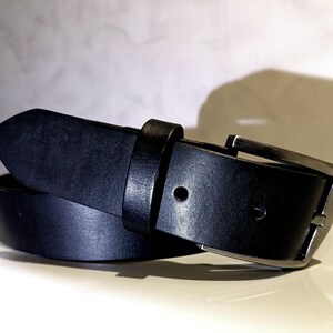 May include: A black leather belt with a silver buckle. The belt is rolled, displaying the leather's texture. The buckle is rectangular with a single prong. The belt's width is approximately 3.8 cm.