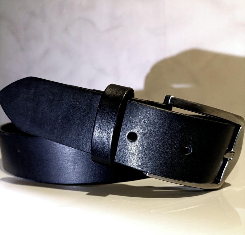 May include: A black leather belt with a silver buckle. The belt is rolled, displaying the leather's texture. The buckle is rectangular with a single prong. The belt's width is approximately 3.8 cm.