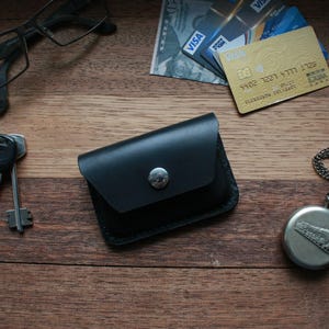 Puede incluir: Una cartera de cuero negro con cierre de presión y un botón plateado. La cartera está sobre una superficie de madera con un par de gafas, llaves y un reloj de bolsillo.