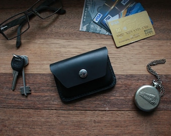 Puede incluir: Una cartera de cuero negro con cierre de presión y un botón plateado. La cartera está sobre una superficie de madera con un par de gafas, llaves y un reloj de bolsillo.