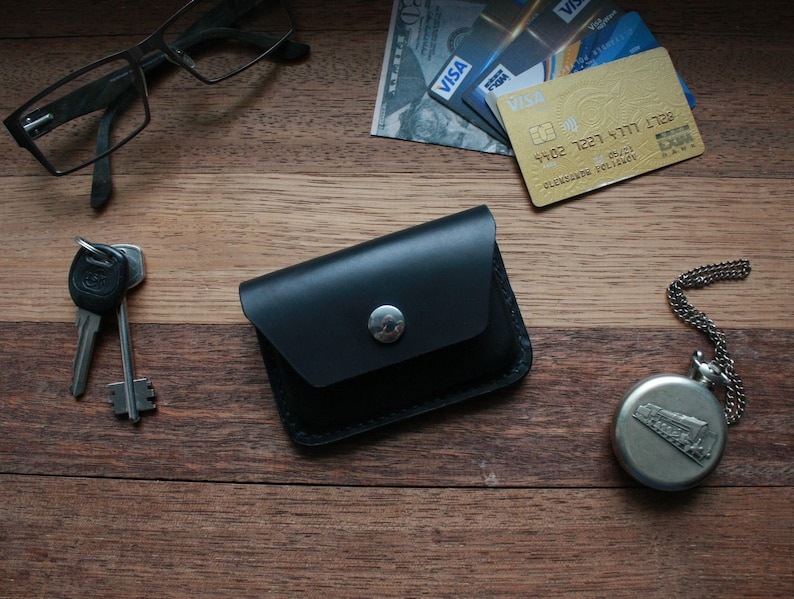 Puede incluir: Una cartera de cuero negro con cierre de presi&oacute;n y un bot&oacute;n plateado. La cartera est&aacute; sobre una superficie de madera con un par de gafas, llaves y un reloj de bolsillo.