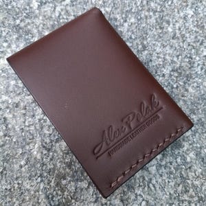 May include: A rectangular dark brown leather card holder featuring visible stitching along the bottom edge. The front surface is embossed with the text Alex Polak and the words HANDMADE LEATHER GOODS positioned directly underneath the brand name.