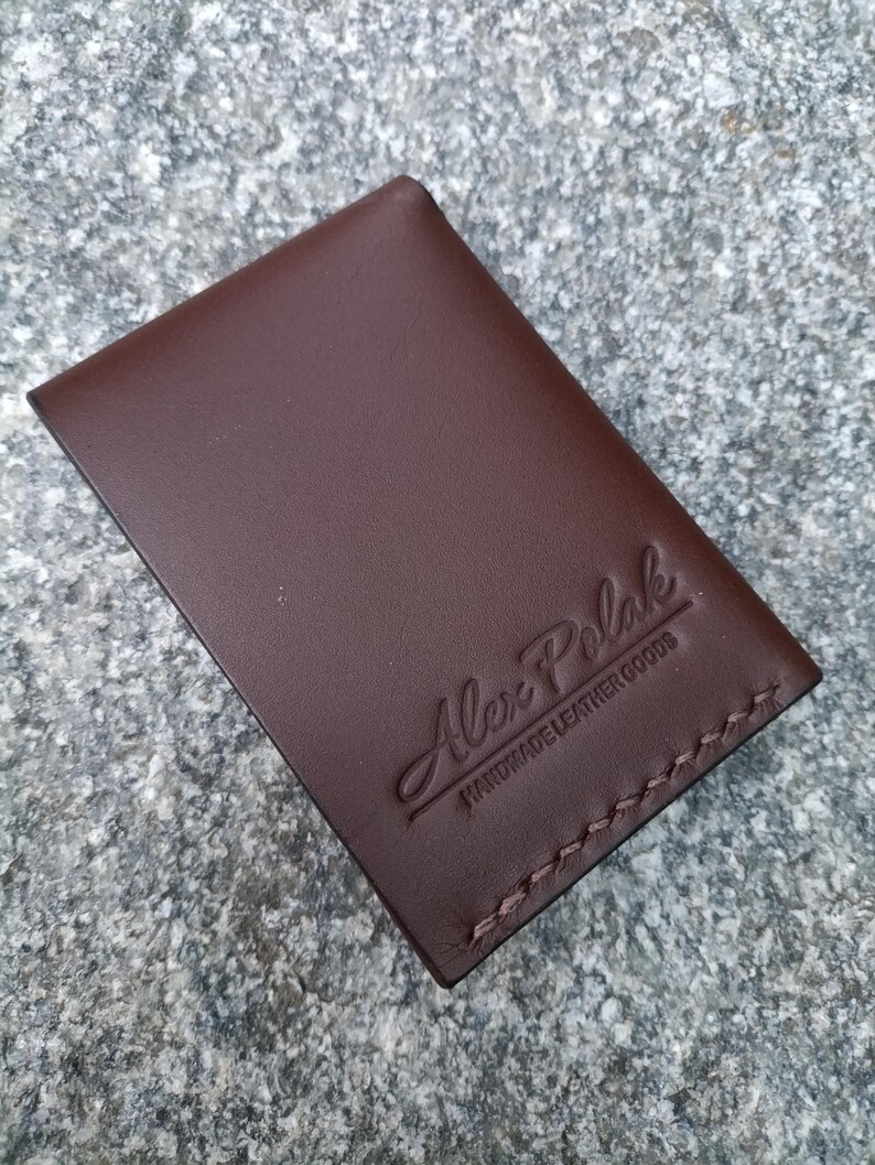 May include: A rectangular dark brown leather card holder featuring visible stitching along the bottom edge. The front surface is embossed with the text Alex Polak and the words HANDMADE LEATHER GOODS positioned directly underneath the brand name.