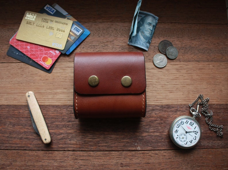 May include: A brown leather wallet with two brass snaps and a pocket watch with a silver chain on a wooden surface.