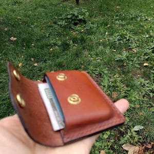 Puede incluir: Una cartera de cuero marr&oacute;n con cierre de solapa asegurado por dos botones de presi&oacute;n de lat&oacute;n. El interior de la cartera es visible, mostrando una ranura para tarjetas que contiene una tarjeta blanca con texto oscuro.