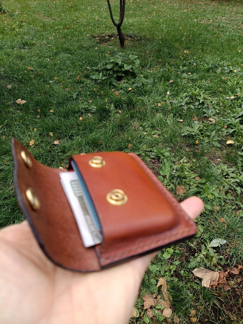 Puede incluir: Una cartera de cuero marr&oacute;n con cierre de solapa asegurado por dos botones de presi&oacute;n de lat&oacute;n. El interior de la cartera es visible, mostrando una ranura para tarjetas que contiene una tarjeta blanca con texto oscuro.
