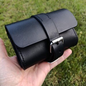May include: A black leather watch roll with a silver buckle closure. The roll is cylindrical and secured with a matching leather strap. The stitching is visible along the edges. The watch roll is held in a hand against a green background.