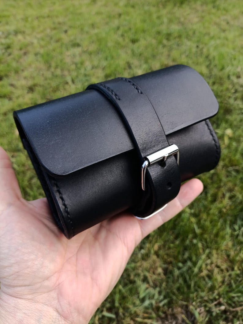 May include: A black leather watch roll with a silver buckle closure. The roll is cylindrical and secured with a matching leather strap. The stitching is visible along the edges. The watch roll is held in a hand against a green background.