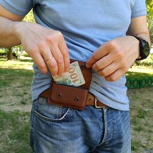 Puede incluir: Una ri&ntilde;onera de cuero marr&oacute;n con cierre de solapa asegurado por dos broches de metal negro. La bolsa est&aacute; sujeta a un cintur&oacute;n de cuero marr&oacute;n y contiene un billete de cien d&oacute;lares doblado parcialmente visible en el compartimento.