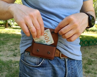 Puede incluir: Una riñonera de cuero marrón con cierre de solapa asegurado por dos broches de metal negro. La bolsa está sujeta a un cinturón de cuero marrón y contiene un billete de cien dólares doblado parcialmente visible en el compartimento.