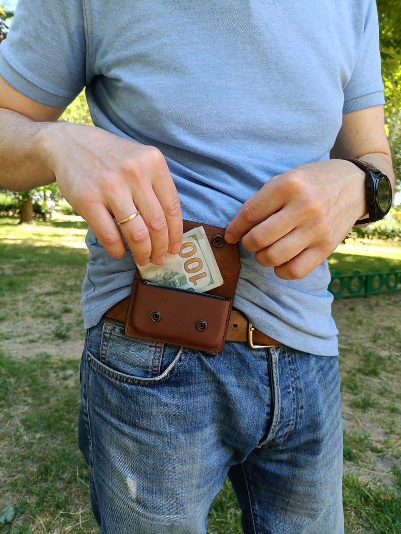 Puede incluir: Una ri&ntilde;onera de cuero marr&oacute;n con cierre de solapa asegurado por dos broches de metal negro. La bolsa est&aacute; sujeta a un cintur&oacute;n de cuero marr&oacute;n y contiene un billete de cien d&oacute;lares doblado parcialmente visible en el compartimento.