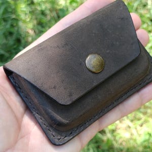 May include: A small, rectangular dark brown leather wallet featuring a smooth, matte finish and a single brass-colored metal snap button closure centered on the front flap. The edges are secured with visible dark stitching along the perimeter.