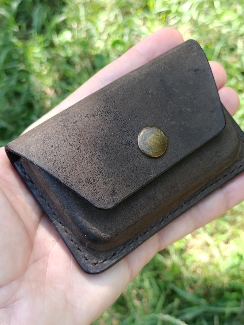May include: A small, rectangular dark brown leather wallet featuring a smooth, matte finish and a single brass-colored metal snap button closure centered on the front flap. The edges are secured with visible dark stitching along the perimeter.