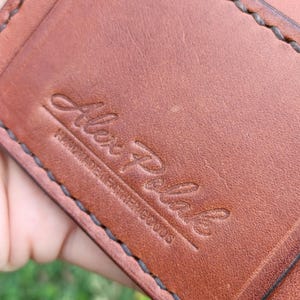 May include: A brown leather wallet featuring dark brown saddle stitching along the edges. The surface is embossed with the script text Alex Polak and the words HANDMADE LEATHER GOODS underneath in a smaller, sans-serif font.