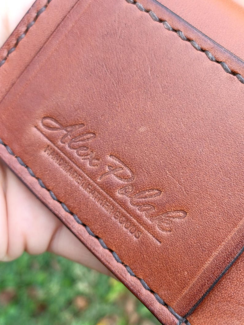 May include: A brown leather wallet featuring dark brown saddle stitching along the edges. The surface is embossed with the script text Alex Polak and the words HANDMADE LEATHER GOODS underneath in a smaller, sans-serif font.