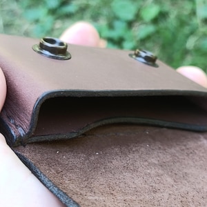 Puede incluir: Una funda de cuero marr&oacute;n con un compartimento abierto y dos cierres de presi&oacute;n de metal oscuro en el borde superior. El interior muestra un acabado de ante texturizado, mientras que el exterior tiene una superficie lisa con costuras visibles.
