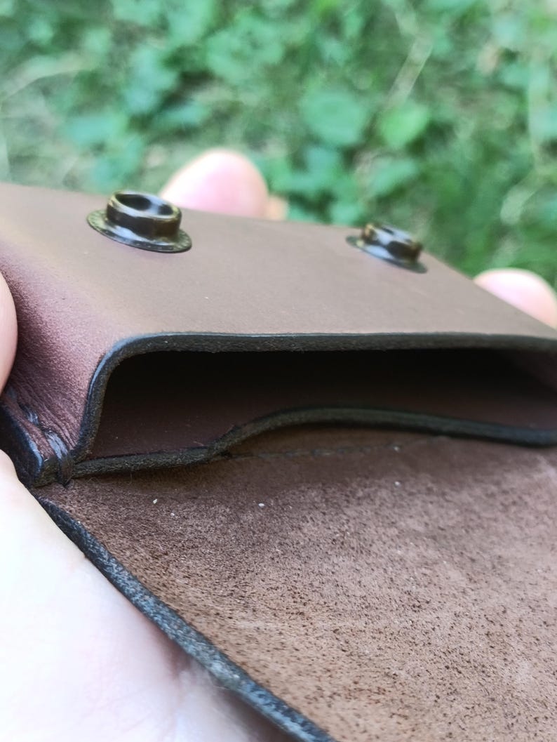 Puede incluir: Una funda de cuero marr&oacute;n con un compartimento abierto y dos cierres de presi&oacute;n de metal oscuro en el borde superior. El interior muestra un acabado de ante texturizado, mientras que el exterior tiene una superficie lisa con costuras visibles.