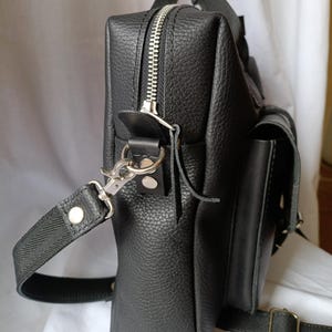 Puede incluir: Bolso mensajero de cuero negro con acabado texturizado. Cuenta con cremallera plateada, asa y correa para el hombro desmontable. Incluye un peque&ntilde;o bolsillo lateral y cierre de hebilla. El bolso mide aproximadamente 25 cm de alto y 20 cm de ancho.