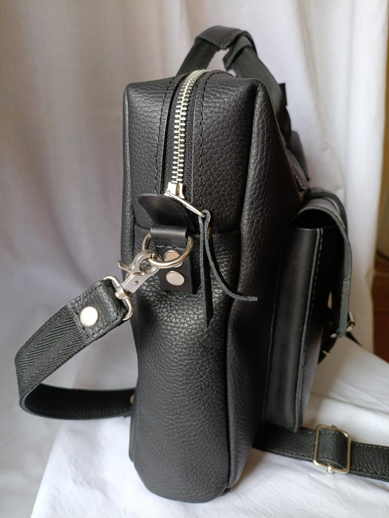 Puede incluir: Bolso mensajero de cuero negro con acabado texturizado. Cuenta con cremallera plateada, asa y correa para el hombro desmontable. Incluye un peque&ntilde;o bolsillo lateral y cierre de hebilla. El bolso mide aproximadamente 25 cm de alto y 20 cm de ancho.