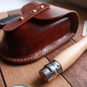 May include: A brown leather pouch with a curved top and a flap secured by copper rivets and a gold stud. A wooden-handled knife with a silver blade is next to the pouch. The items are on a wooden surface.