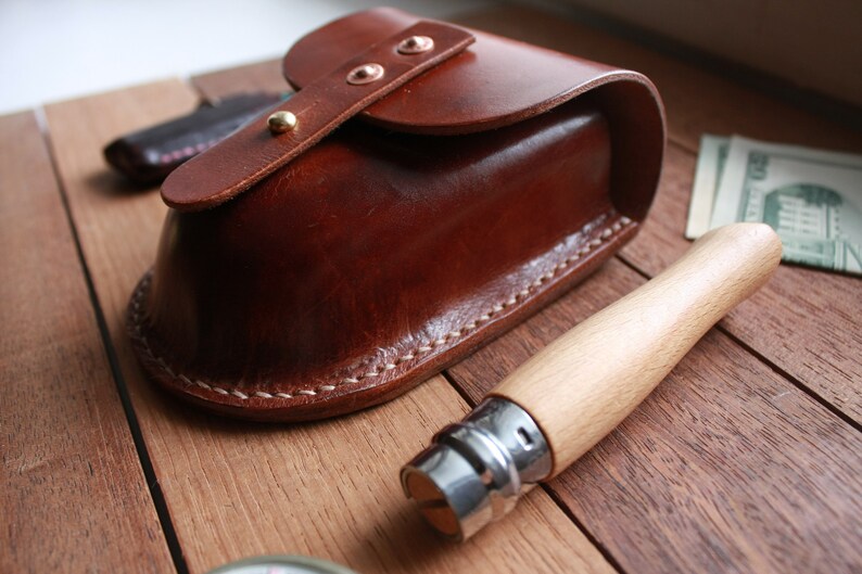 May include: A brown leather pouch with a curved top and a flap secured by copper rivets and a gold stud. A wooden-handled knife with a silver blade is next to the pouch. The items are on a wooden surface.