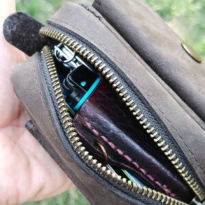Peut inclure: Une pochette en cuir marron fonc&eacute; avec une fermeture &eacute;clair en m&eacute;tal couleur laiton. La pochette est entrouverte, r&eacute;v&eacute;lant un objet en cuir violet fonc&eacute; avec des coutures roses et une partie d'un appareil &eacute;lectronique noir et bleu sarcelle.