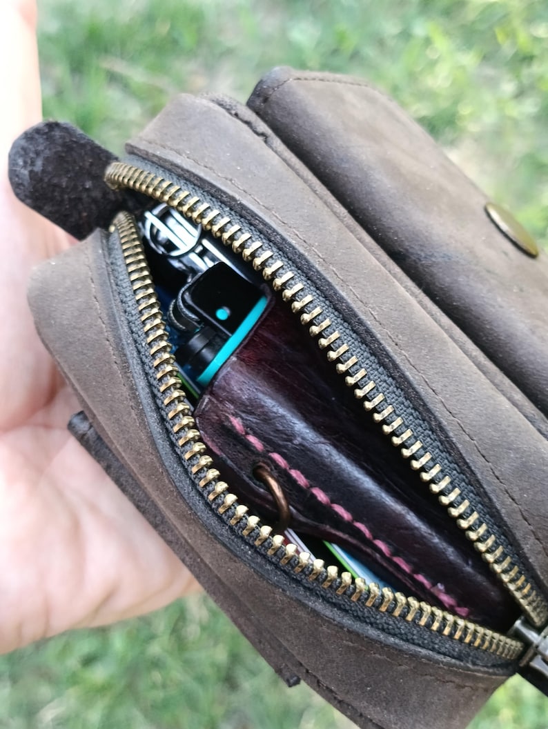 Peut inclure: Une pochette en cuir marron fonc&eacute; avec une fermeture &eacute;clair en m&eacute;tal couleur laiton. La pochette est entrouverte, r&eacute;v&eacute;lant un objet en cuir violet fonc&eacute; avec des coutures roses et une partie d'un appareil &eacute;lectronique noir et bleu sarcelle.