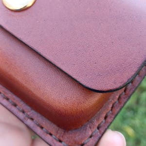 May include: A brown leather pouch featuring a smooth, rounded flap secured by a polished gold-tone metal snap button. The edges of the pouch are joined with thick, dark brown stitching, showcasing a sturdy construction and a natural grain texture.