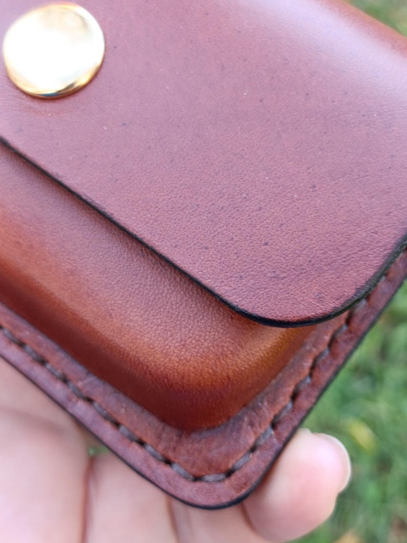 May include: A brown leather pouch featuring a smooth, rounded flap secured by a polished gold-tone metal snap button. The edges of the pouch are joined with thick, dark brown stitching, showcasing a sturdy construction and a natural grain texture.