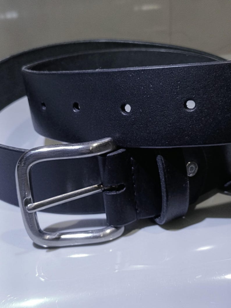 Op de afbeelding: Een zwarte leren riem met een zilverkleurige gesp. De riem heeft verschillende verstelgaten en een klassiek, minimalistisch ontwerp. De rechthoekige gesp heeft een enkele pin.
