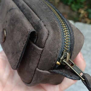 Peut inclure: Une pochette en daim marron fonc&eacute; dot&eacute;e d'un compartiment principal zipp&eacute; avec une tirette en m&eacute;tal couleur laiton et d'une petite poche avant s&eacute;curis&eacute;e par un bouton-pression rond assorti, pr&eacute;sentant une finition mate textur&eacute;e.