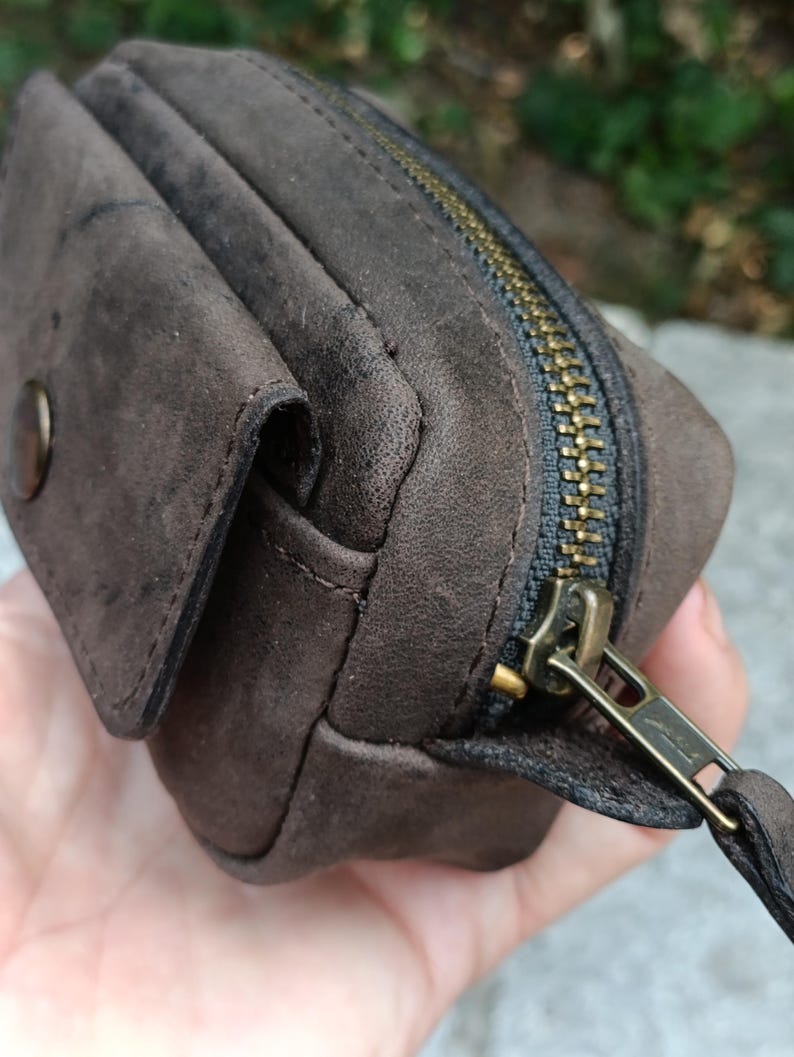 Peut inclure: Une pochette en daim marron fonc&eacute; dot&eacute;e d'un compartiment principal zipp&eacute; avec une tirette en m&eacute;tal couleur laiton et d'une petite poche avant s&eacute;curis&eacute;e par un bouton-pression rond assorti, pr&eacute;sentant une finition mate textur&eacute;e.