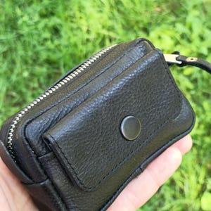 Puede incluir: Un peque&ntilde;o estuche de cuero negro texturizado con un compartimento principal con cremallera y un bolsillo frontal asegurado con un bot&oacute;n a presi&oacute;n negro mate. El accesorio rectangular incluye una correa de mu&ntilde;eca de cuero negro.