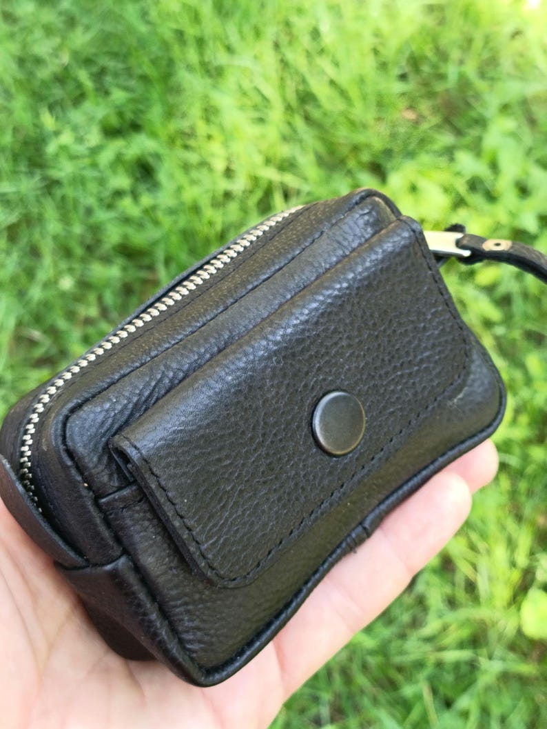 Puede incluir: Un peque&ntilde;o estuche de cuero negro texturizado con un compartimento principal con cremallera y un bolsillo frontal asegurado con un bot&oacute;n a presi&oacute;n negro mate. El accesorio rectangular incluye una correa de mu&ntilde;eca de cuero negro.