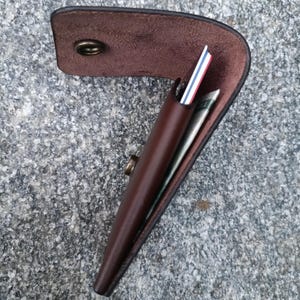 May include: A dark brown leather wallet with a curved flap and a brass snap button closure. The wallet is partially open, revealing a stack of cards with white, red, and blue edges, along with a folded paper bill tucked inside the main compartment.