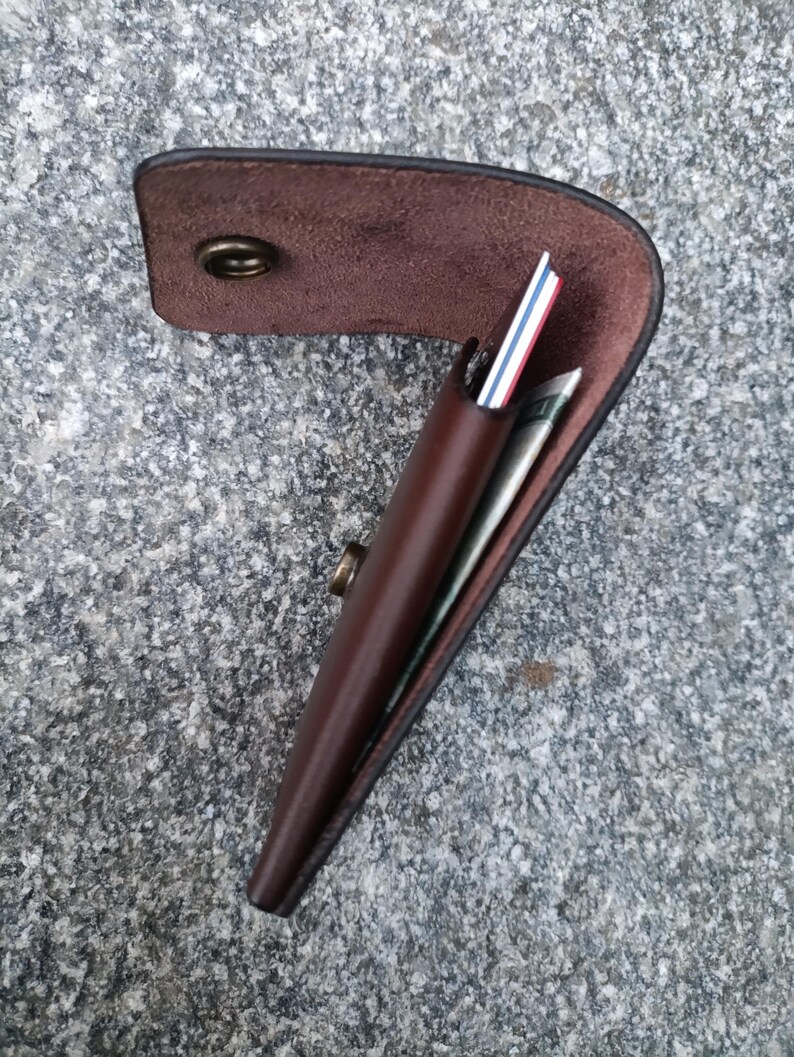 May include: A dark brown leather wallet with a curved flap and a brass snap button closure. The wallet is partially open, revealing a stack of cards with white, red, and blue edges, along with a folded paper bill tucked inside the main compartment.