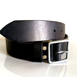以下が含まれることがあります： シルバーのバックルが付いた黒い革ベルト。 長方形のバックルと2つのシルバーリベットを備えたクラシックなデザインです。 革はわずかに使い古された仕上がりになっています。 ベルトの幅は約3.8cmです。
