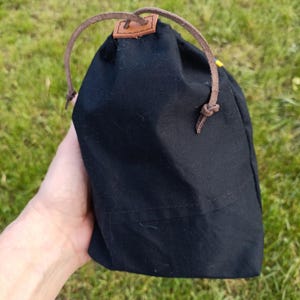 May include: A black drawstring bag with a brown leather strap and a small brown leather patch. The bag is made of a dark fabric and is held against a background of green grass.
