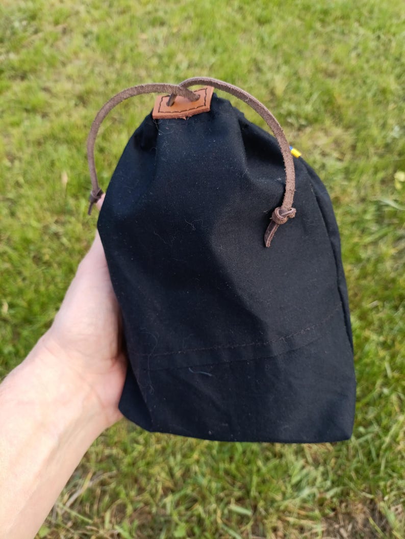 May include: A black drawstring bag with a brown leather strap and a small brown leather patch. The bag is made of a dark fabric and is held against a background of green grass.