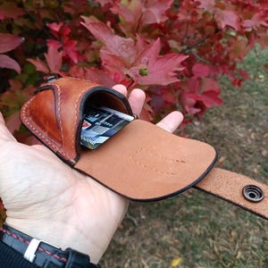 K&ouml;nnte beinhalten: Ein brauner Lederkartenhalter mit Druckknopfverschluss und G&uuml;rtelschlaufe. Der Kartenhalter ist ge&ouml;ffnet und zeigt einige Karten im Inneren. Der Hintergrund zeigt rote und gr&uuml;ne Bl&auml;tter.
