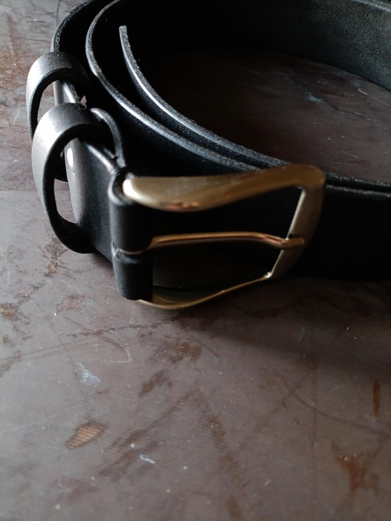 Puede incluir: Un cintur&oacute;n negro con un acabado liso y mate y una hebilla de metal en tono lat&oacute;n pulido. El cintur&oacute;n incluye dos trabillas de cuero negro a juego cerca de la hebilla, la cual est&aacute; sujeta a la tira con un remache met&aacute;lico visible.