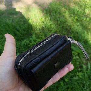 Puede incluir: Una peque&ntilde;a bolsa de cuero negro texturizado con una cremallera de metal plateado en la parte superior y un bolsillo frontal asegurado con un bot&oacute;n a presi&oacute;n negro a juego. El accesorio rectangular tiene un dise&ntilde;o compacto y costuras.