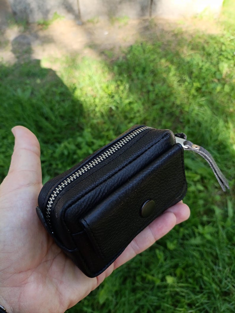 Puede incluir: Una peque&ntilde;a bolsa de cuero negro texturizado con una cremallera de metal plateado en la parte superior y un bolsillo frontal asegurado con un bot&oacute;n a presi&oacute;n negro a juego. El accesorio rectangular tiene un dise&ntilde;o compacto y costuras.