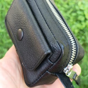Puede incluir: Una peque&ntilde;a bolsa negra texturizada que cuenta con una solapa frontal con un cierre de bot&oacute;n a presi&oacute;n y una cremallera de metal plateado en el borde superior. El accesorio rectangular tiene costuras visibles y una leng&uuml;eta de cuero.