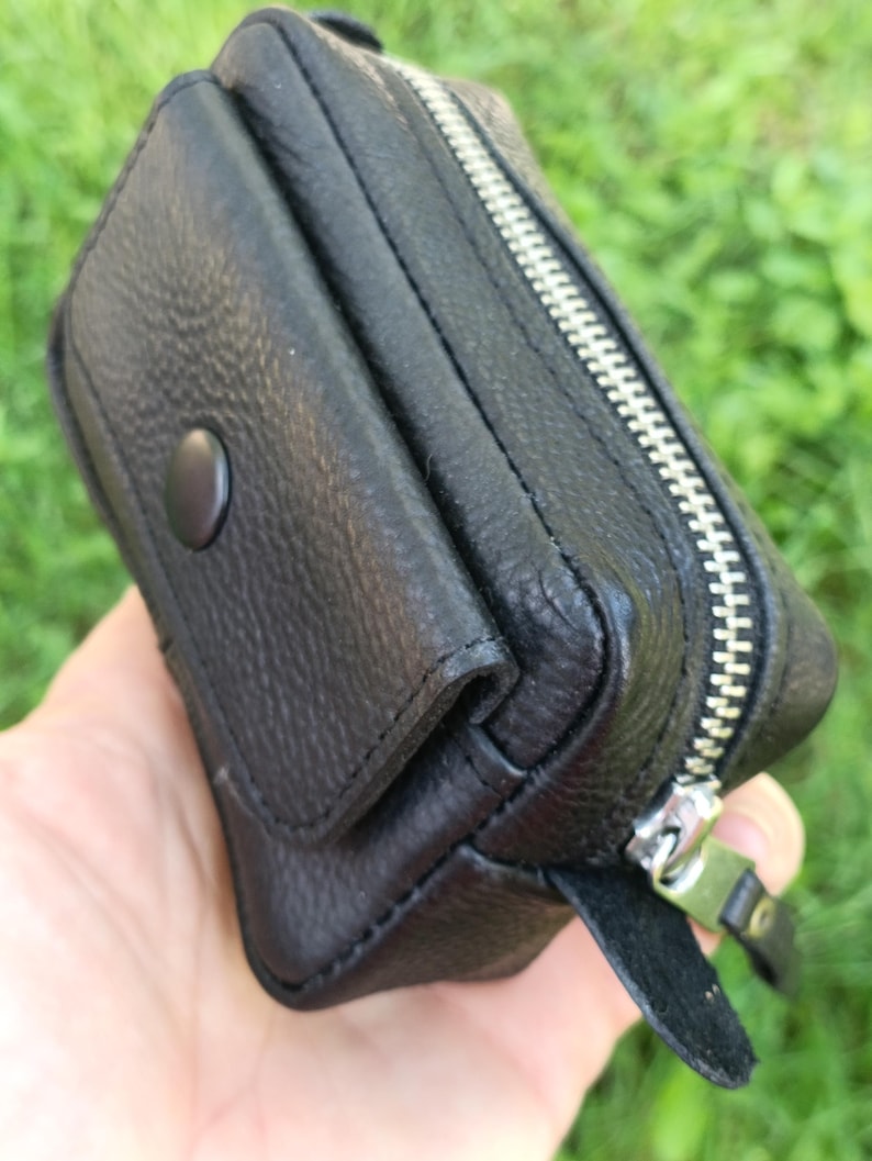 Puede incluir: Una peque&ntilde;a bolsa negra texturizada que cuenta con una solapa frontal con un cierre de bot&oacute;n a presi&oacute;n y una cremallera de metal plateado en el borde superior. El accesorio rectangular tiene costuras visibles y una leng&uuml;eta de cuero.