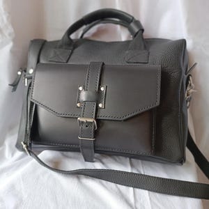 Puede incluir: Un bolso de cuero negro con asa superior y bandolera desmontable. El bolso presenta un bolsillo frontal con solapa asegurado con un cierre de hebilla y herrajes plateados. El cuero tiene un grano texturizado.