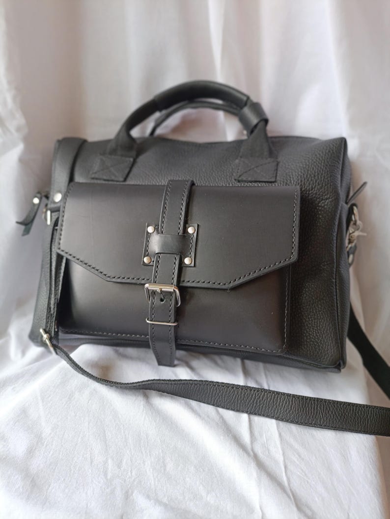 Puede incluir: Un bolso de cuero negro con asa superior y bandolera desmontable. El bolso presenta un bolsillo frontal con solapa asegurado con un cierre de hebilla y herrajes plateados. El cuero tiene un grano texturizado.