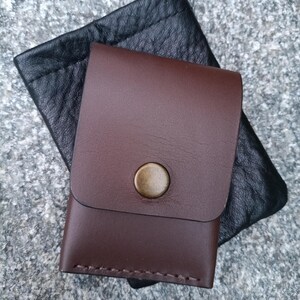 May include: A smooth dark brown leather card holder with a single brass snap button closure, featuring visible stitching along the bottom edge, layered over a black textured leather pouch with a drawstring top closure.